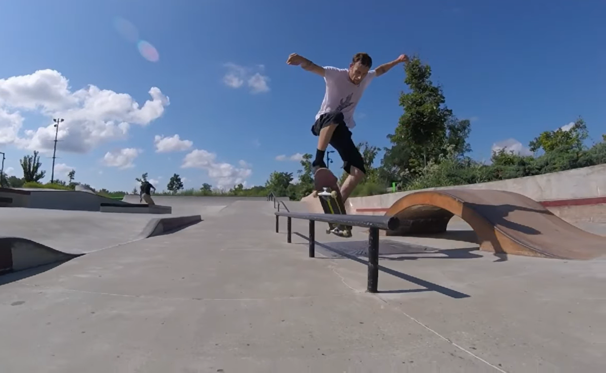 A man in a white t-shirt and black shorts grinds a skateboard across a rail at Bloomington's Switchyard skatepark.