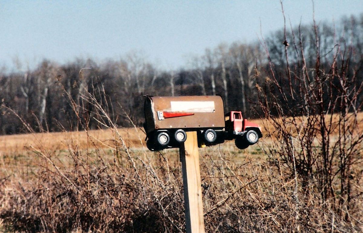 a mailbox decorated like a tractor-trailer on a wooden post before a background of leafless trees and a brown field