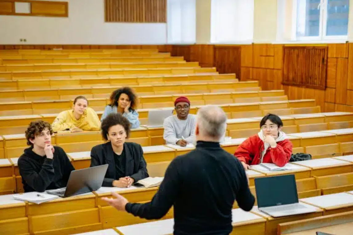 a man speaks to a diverse group of six students in a college classroom setting