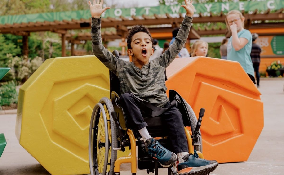 a young boy in a wheelchair exclaims with his arms in the air in front of a brightly colored sculpture spelling "ZOO"
