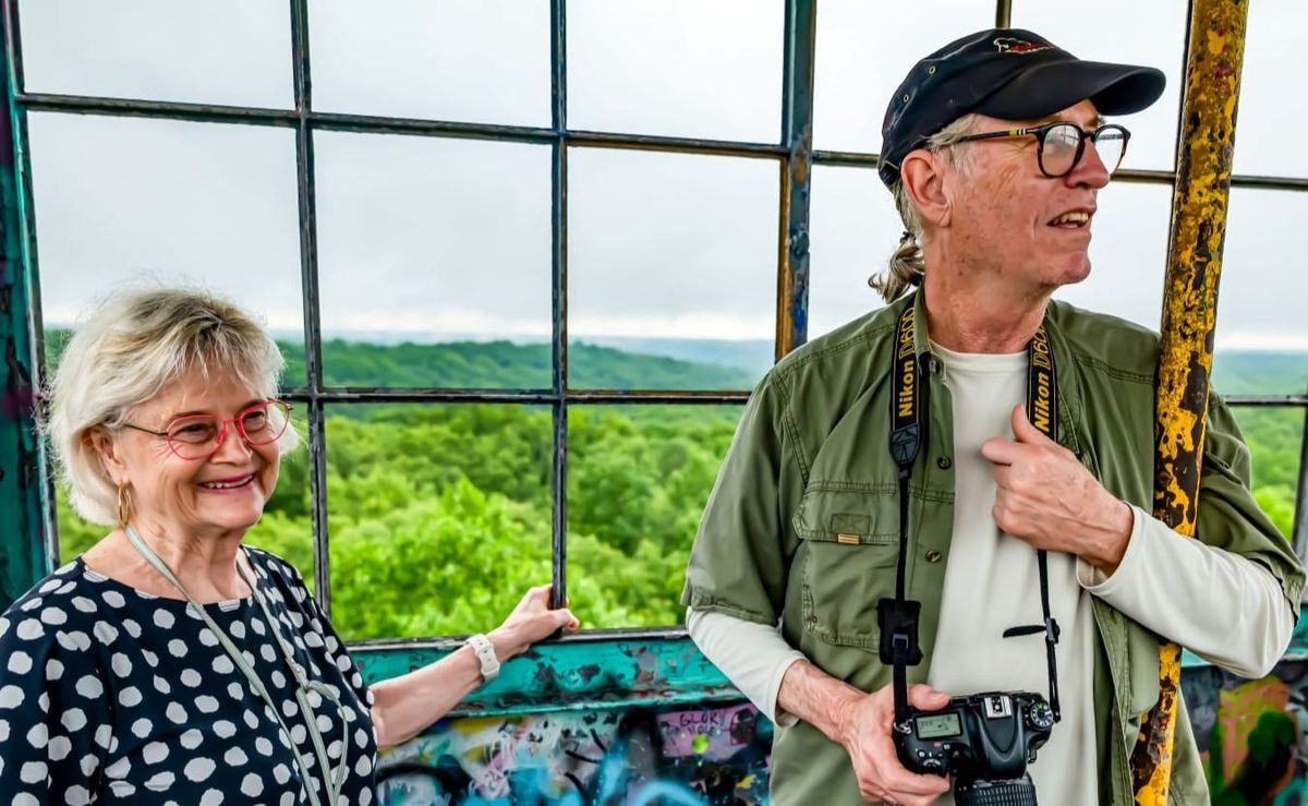 a woman in red glasses and black and white polka-dot shirt stands next to a man in black cap, black glasses, and green shirt over tan undershirt, inside a fire tower, green trees below