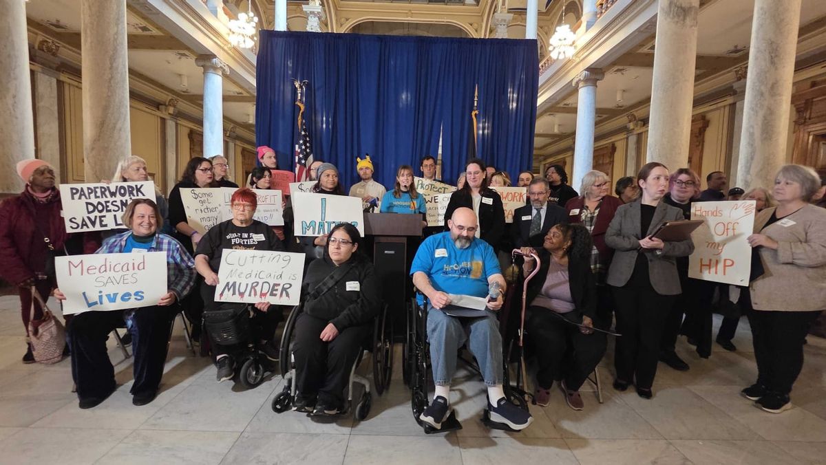 In an ornate room with stone columns a woman speaks at a lectern in front of a blue curtain. A surrounding group of people, some in wheelchairs, hold signs promoting accessible healthcare