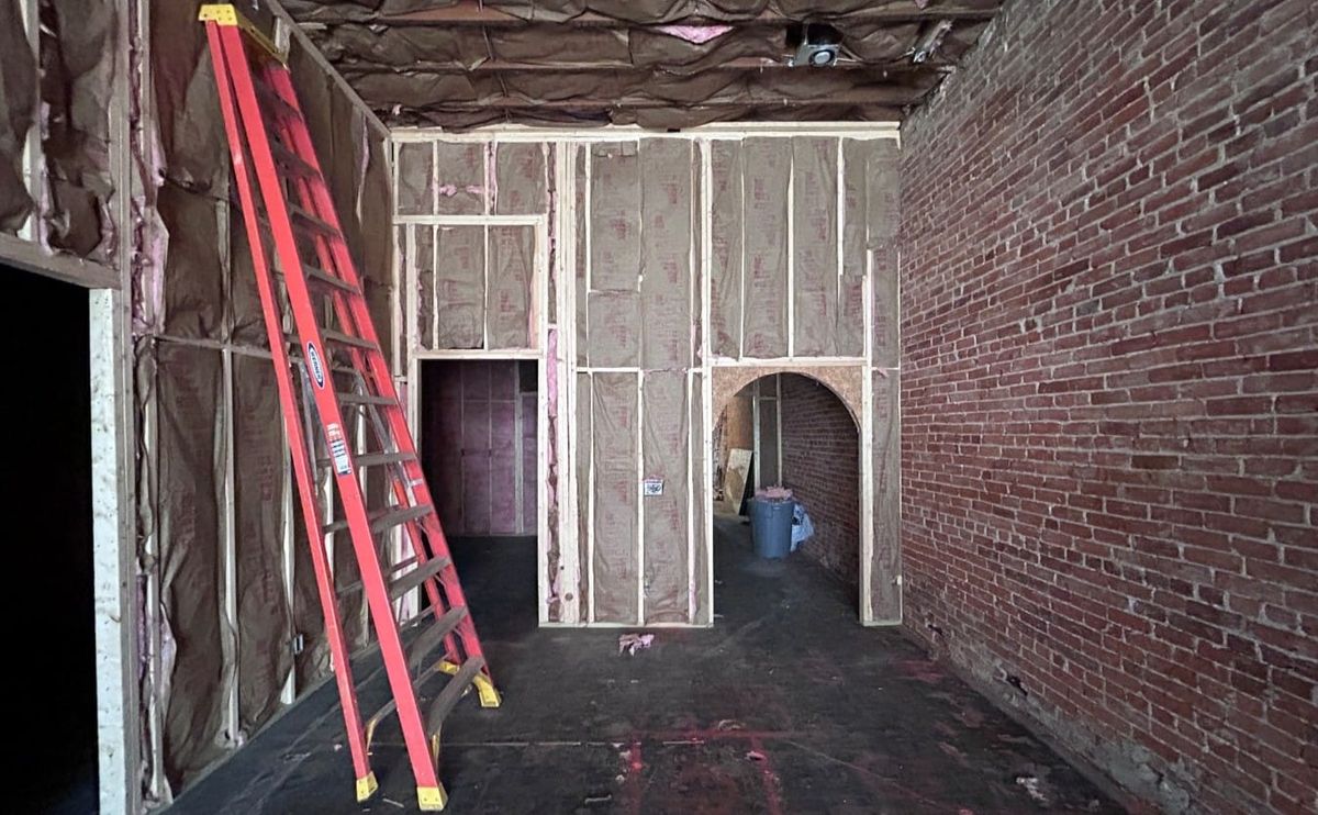 the interior of a building. A brick wall adjoins bare wood framed walls stuffed with insulation. A ladder leans against the constrcution