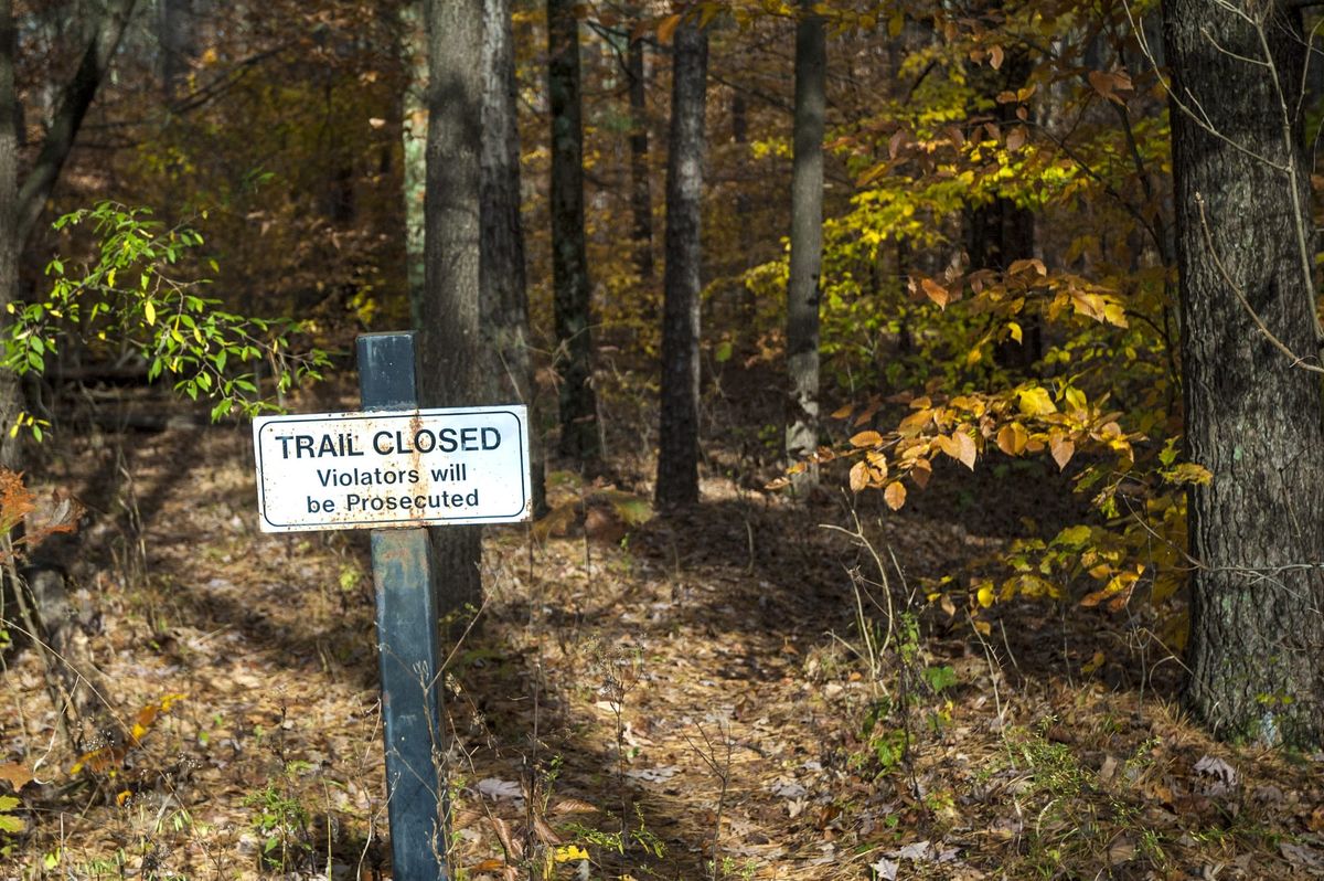 a metal sign on a metal post in a brown forest with autumn colors. The sign reads "TRAIL CLOSED Violators will be Prosectured"