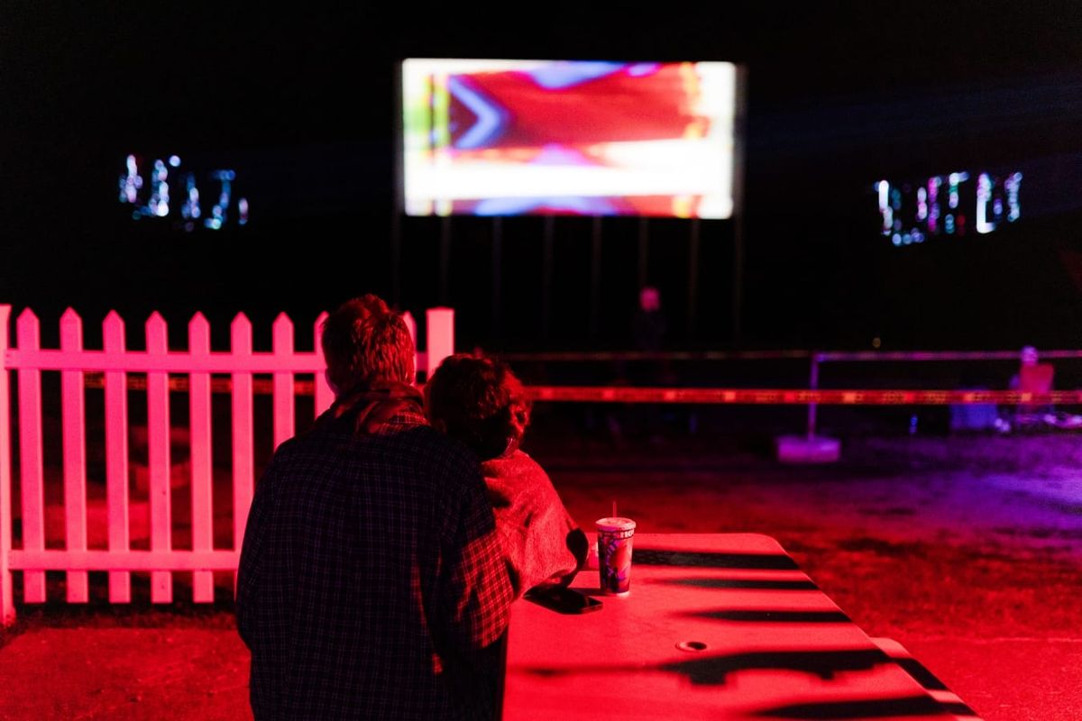 illuminated in red, a man and a woman sit at a table in front of a white picket fence, watching the screen of a drive-in theater. Their backs are toward the camera; a paper cup on the table