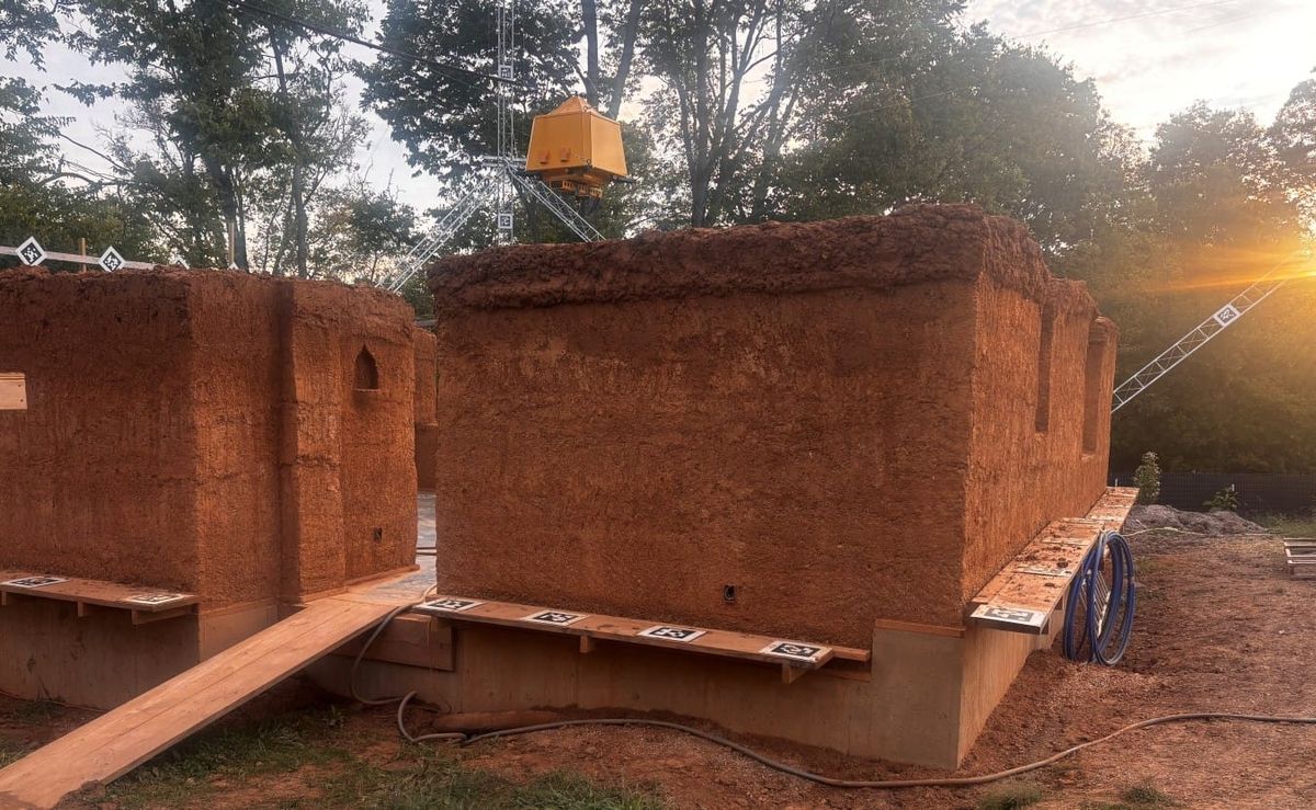 a cob wall house being built with help from a robotic claw device suspended from cables