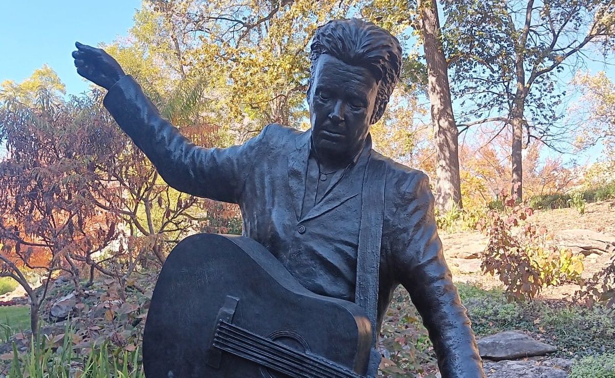 a bronze statue of John Mellencamp holding a guitar, his right arm extended into the air. The background is various autumn foliage