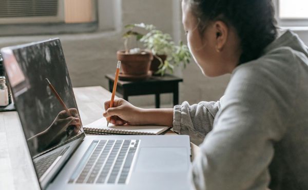 A teenage girl writes with a pencil in a notebook while sitting at a desk, a laptop open in front of her.