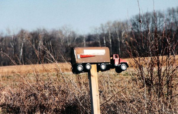 a mailbox decorated like a tractor-trailer on a wooden post before a background of leafless trees and a brown field