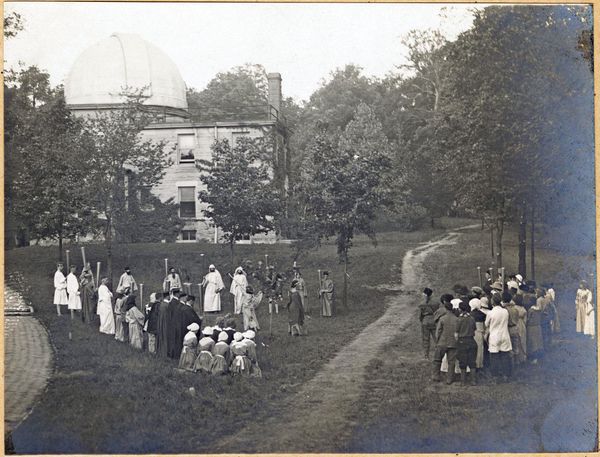 a sepia-toned photograph depicting a group of people in costume performing in a field of grass; an observatory building looms above a few small trees in the background