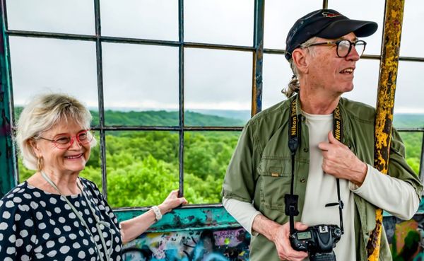 a woman in red glasses and black and white polka-dot shirt stands next to a man in black cap, black glasses, and green shirt over tan undershirt, inside a fire tower, green trees below