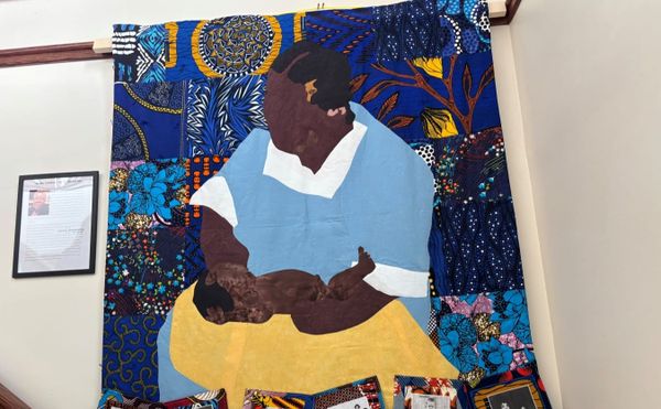 a quilt depicting a Black woman in a blue and white dress holding a Black baby and a yellow blanket. Blue nature motifs make up the background
