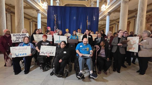 In an ornate room with stone columns a woman speaks at a lectern in front of a blue curtain. A surrounding group of people, some in wheelchairs, hold signs promoting accessible healthcare