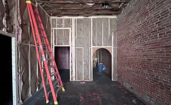 the interior of a building. A brick wall adjoins bare wood framed walls stuffed with insulation. A ladder leans against the constrcution