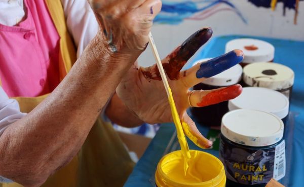 close up of woman's hands as she stirs yellow paint. The fingers of her left hand are each painted a different color: brown, blue, red, yellow