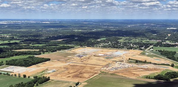 aerial photo of a large construction site in a forested, rural area. Houses and fields are visible under a vast blue sky. Indianapolis skyline on the horizon