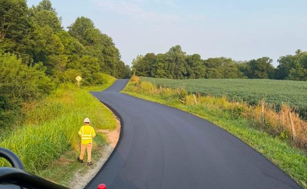 a man wearing high-visibility construction clothes stands in the grass beside a newly paved rural road