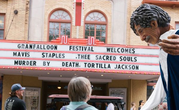 A marquee attached to a yellow brick building announces acts at the Granfalloon Festival. A crowd interacts with a large puppet of Kurt Vonnegut outside