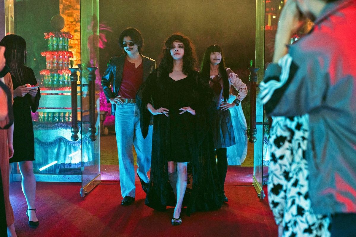 From left to right: a man wearing sunglasses, jeans, a leather jacket and a red shirt, a woman in a black dress with smeared makeup, and a woman wearing a school girl outfit enter a party.