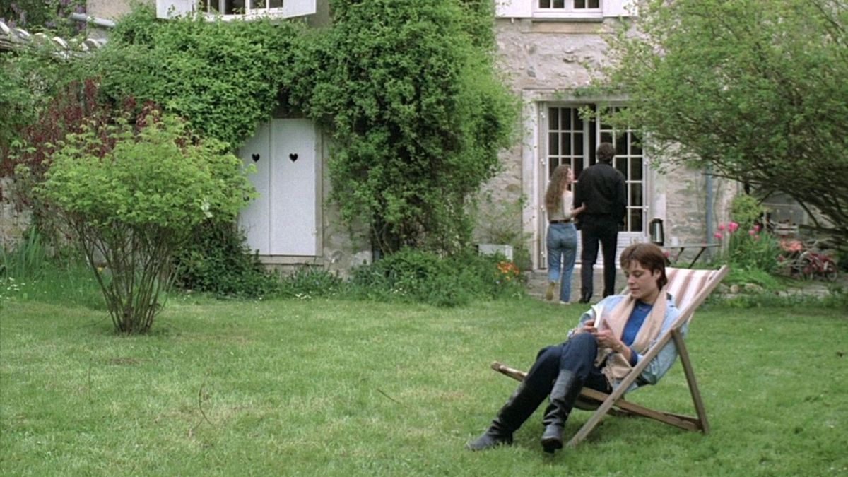 In the foreground, a white woman with cropped hair reads a book in a lounger next to a large house. Behind her, a young woman holds an older man’s arm as they talk, backs to the camera.