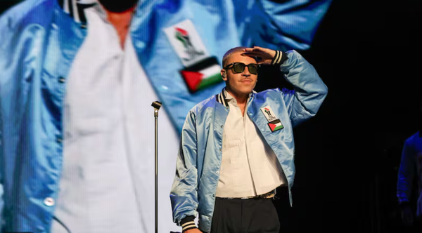 The rapper Macklemore stands on stage saluting. He wears a blue satin jacket with Palestinian flag emblems, a white button-down shirt, and black sunglasses.