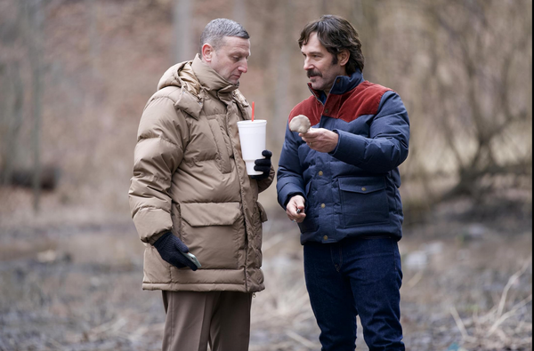 Two middle-aged men stand in a forest clearing. One holds a Big Gulp and the other a mushroom.