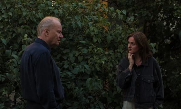 A middle-aged man with grey hair wearing a blue shirt and a thirtysomething woman with shoulder-length brown hair wearing a denim jacket stand in front of a hedge at dusk.