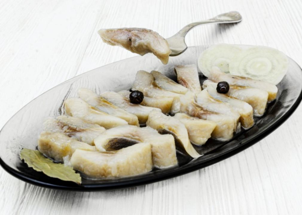 Pickled herring A glass serving dish filled with neatly arranged slices of pickled herring, garnished with onion slices and black peppercorns.