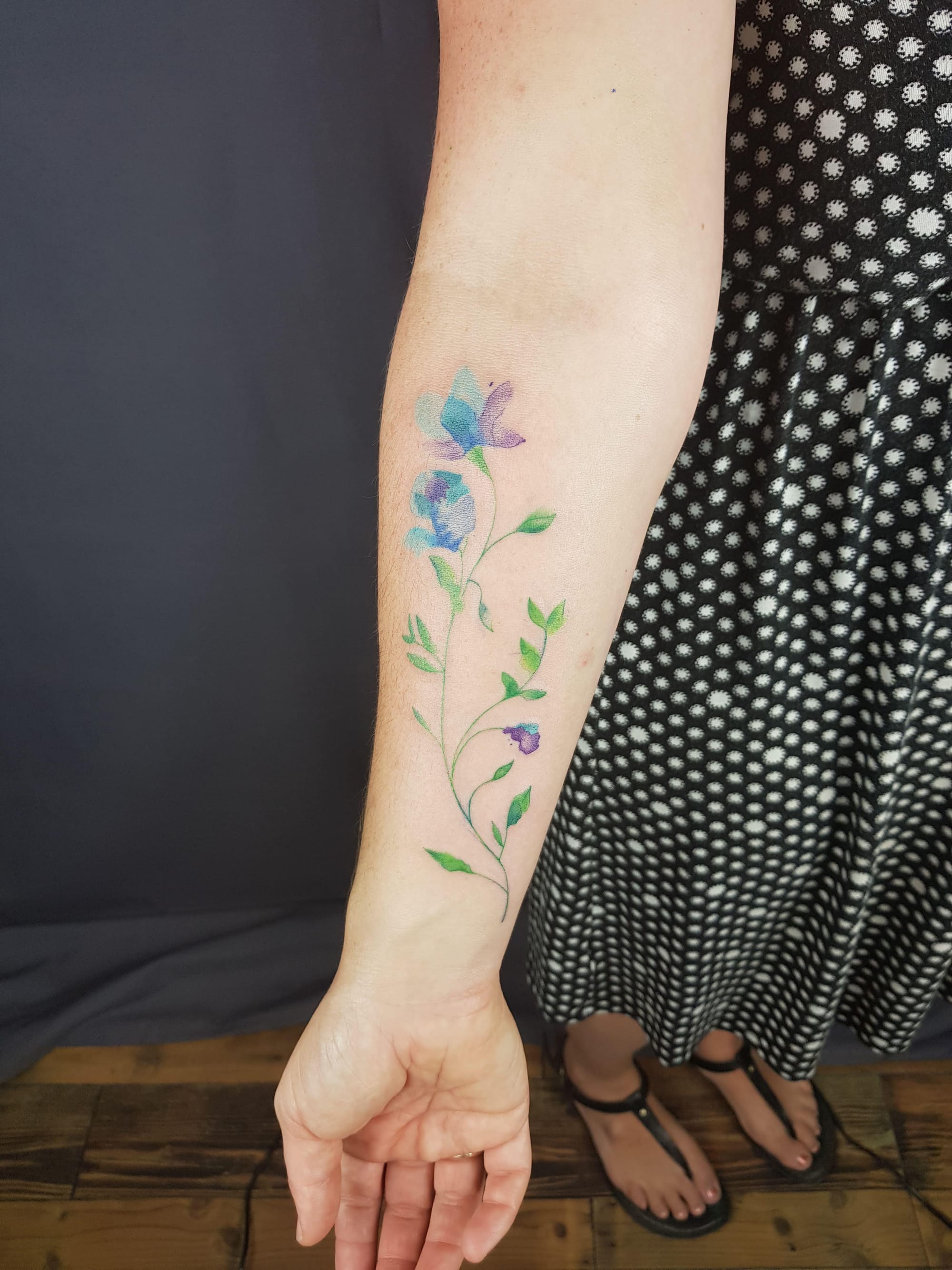 Watercolor magic blooms along this forearm in the most enchanting floral vine tattoo 🌸 The delicate blue and purple petals seem to dance with an almost translucent quality, while graceful green stems wind naturally down the arm like nature's own masterpiece. Perfect placement for a design that flows beautifully with the body's natural lines!