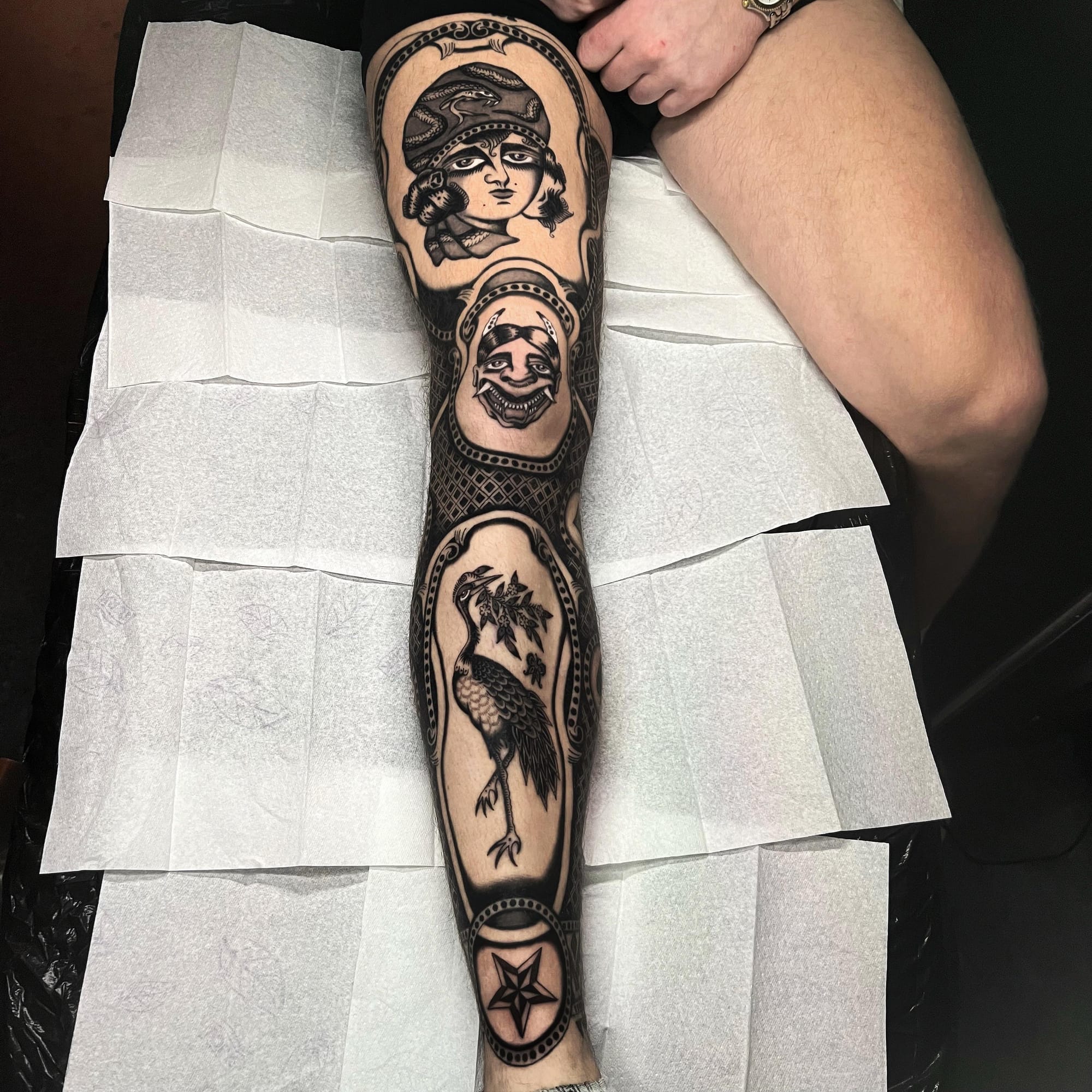 Bold traditional imagery cascades down this stunning leg sleeve, featuring classical portraits framed by intricate geometric borders that create a museum-worthy display. The artist's masterful use of negative space between each vignette allows the woman's face, grotesque mask, and perched raven to each command attention while flowing seamlessly together 🖤 A sophisticated approach to traditional tattooing that proves black and grey work can be just as striking as full color.