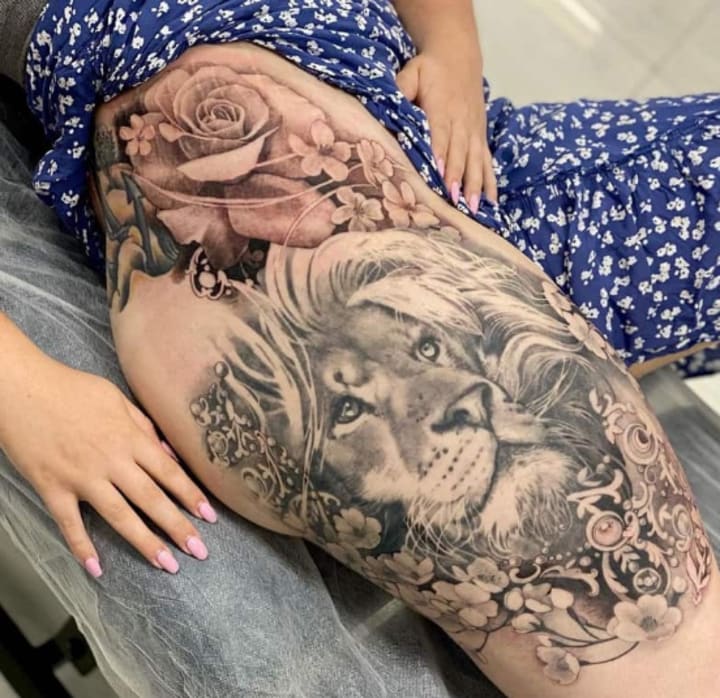 Majestic realism meets feminine artistry in this breathtaking thigh piece that seamlessly blends a powerful lion's gaze with cascading roses and delicate florals. The artist's masterful shading technique creates incredible depth, making the lion's eyes appear almost alive while the botanical elements flow organically around the composition. This is the kind of large-scale tattoo that transforms the leg into a living canvas of strength and beauty ✨