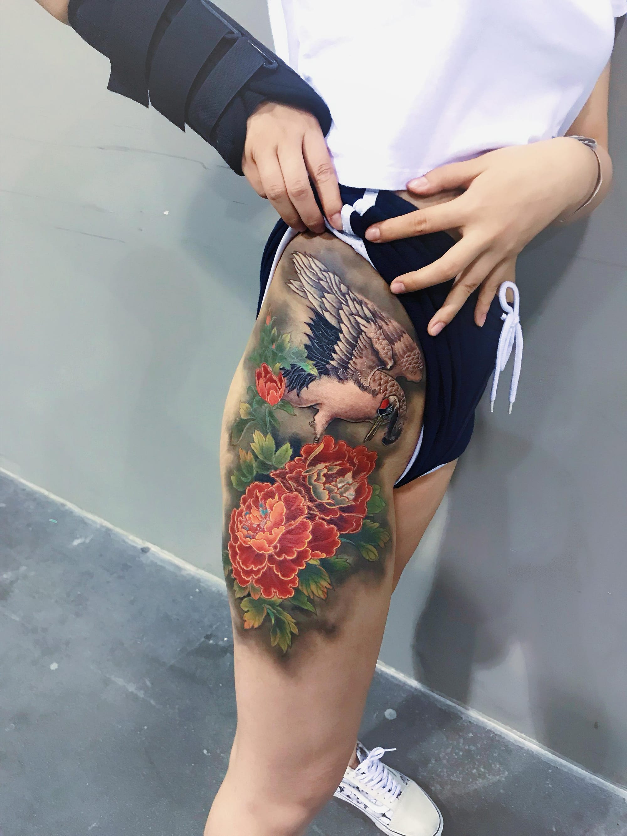 Graceful wings spread across this stunning thigh piece, where a majestic crane takes center stage among blooming peonies in rich coral and crimson hues. The traditional Asian artistry shines through every delicate feather and petal, creating a harmonious balance between the bird's elegant pose and the lush floral backdrop 🌸 This placement perfectly showcases the tattoo's grand scale and intricate details.