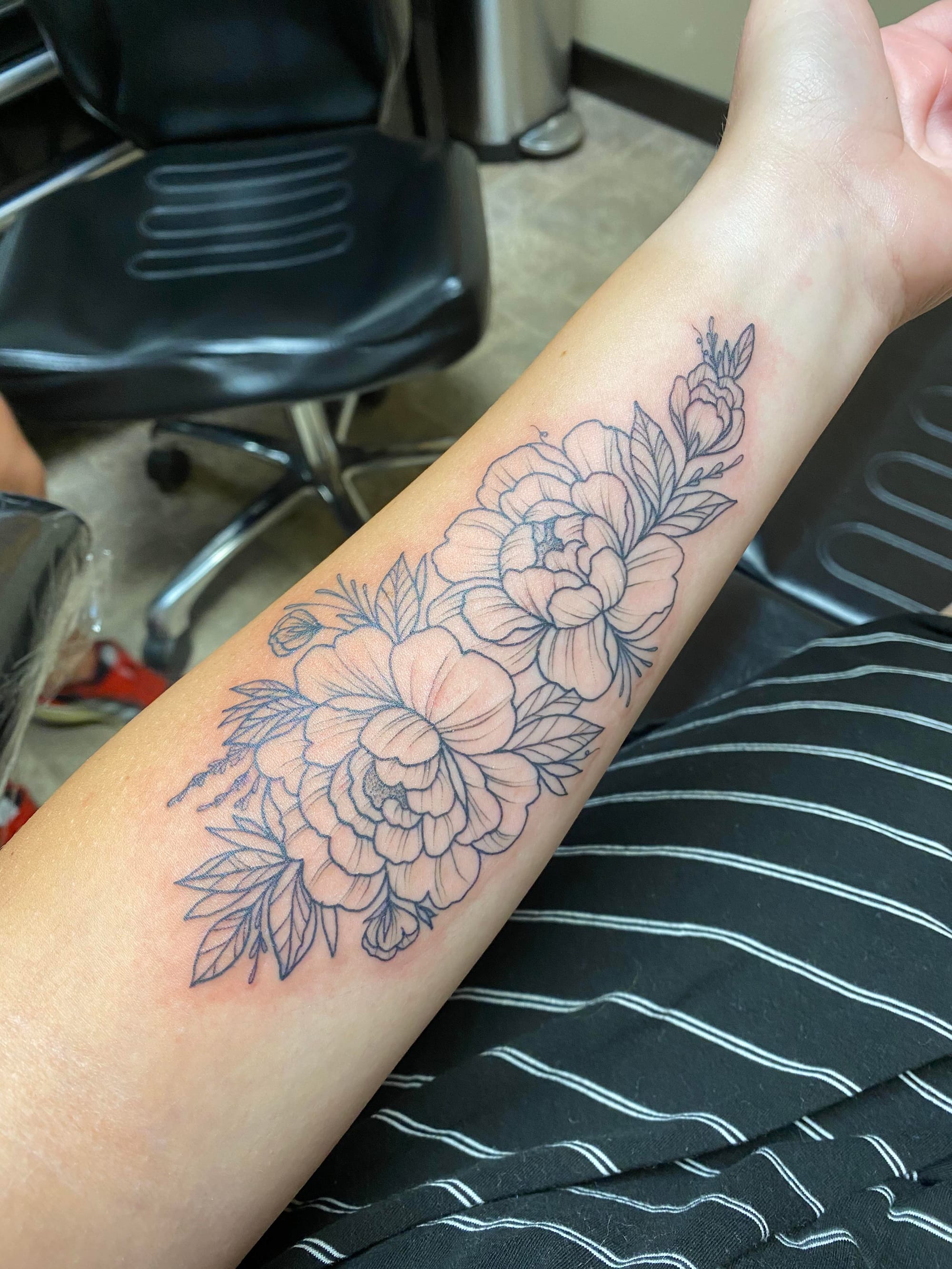 Delicate line work transforms this forearm into a botanical masterpiece, where lush peonies cascade in perfect harmony with intricate leaves and stems. The monochromatic design showcases exceptional shading technique, creating depth and dimension that makes each petal appear to unfold naturally on the skin. A timeless piece that celebrates the elegant beauty of floral artistry 🌸