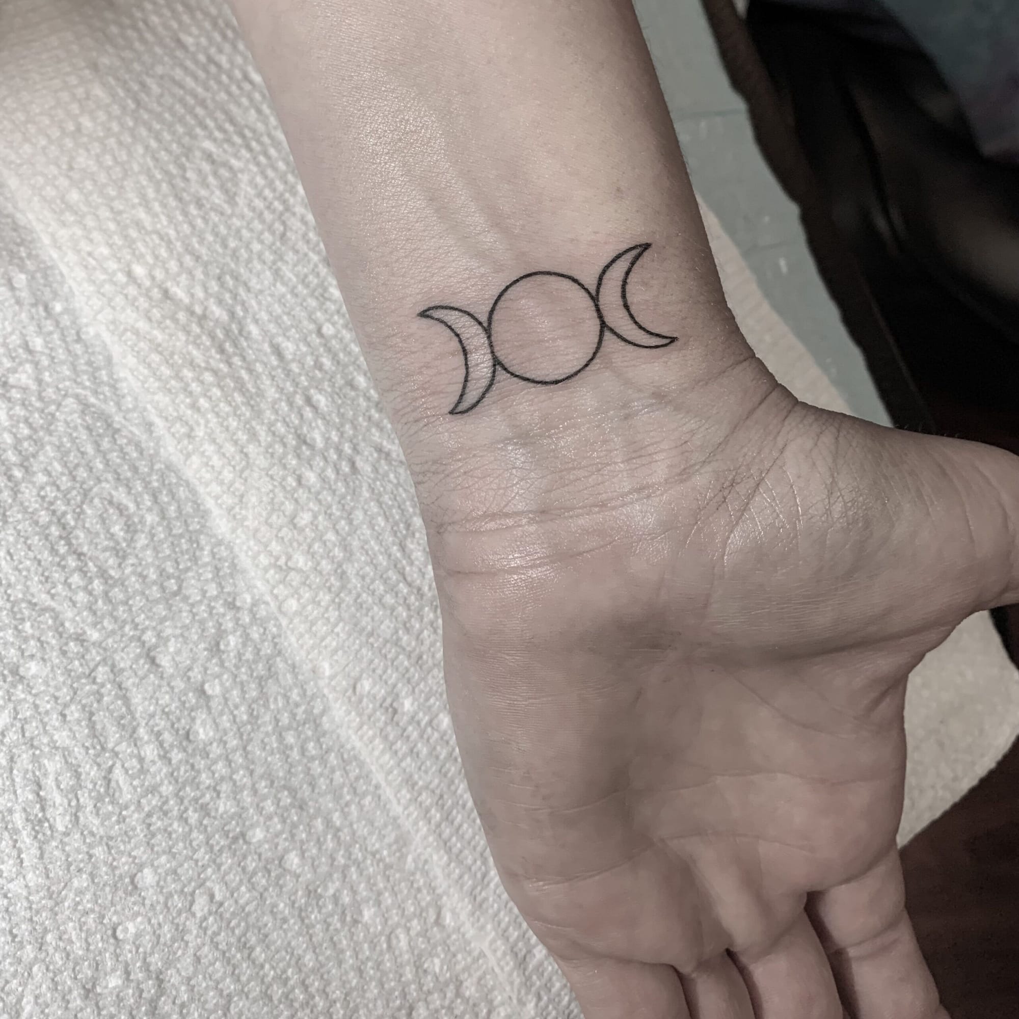 Delicate linework transforms the wrist into a celestial canvas with this minimalist triple moon symbol, representing the maiden, mother, and crone phases of feminine energy. The fine black ink and precise placement create an elegant piece that flows beautifully with the natural contours of the hand and wrist. ✨