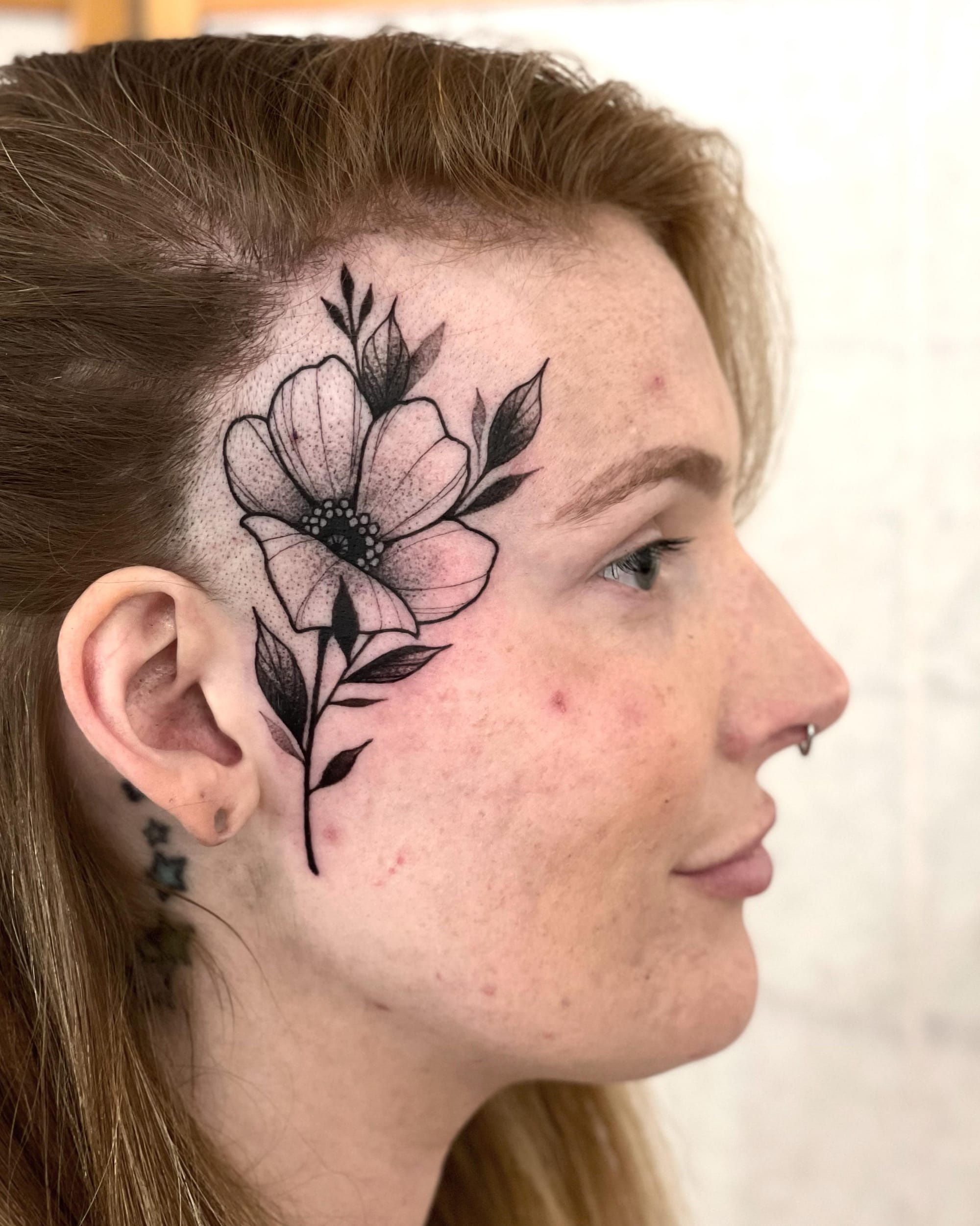 Delicate blackwork blooms across this stunning temple placement, where botanical artistry meets bold placement choices. The intricate dotwork petals and flowing stem create a garden that frames the face beautifully, proving that floral tattoos can be both soft and striking 🌸
