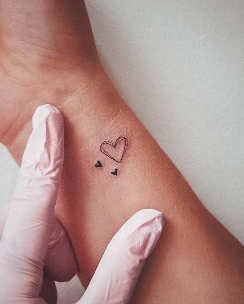 Delicate line work creates pure magic with this minimalist heart design, perfectly positioned on the inner wrist where every glance becomes a gentle reminder of love. The tiny scattered elements add an whimsical touch that elevates this simple concept into something truly special ✨ Fine line tattoos like this prove that sometimes the smallest gestures make the biggest impact.