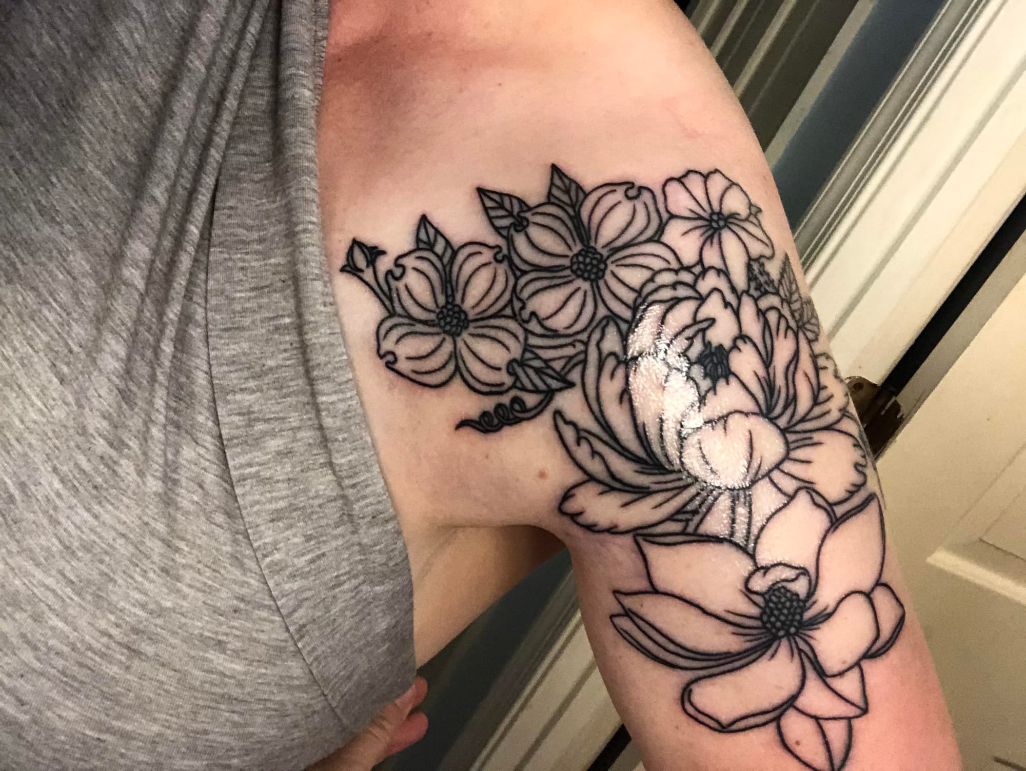 Delicate linework transforms this upper arm into a blooming garden sanctuary, where peonies and botanical beauties cascade in perfect harmony. The masterful black ink outline creates stunning depth through varied petal textures—from smooth, flowing curves to intricate stippled centers that catch the light beautifully. 🌸 Pure floral artistry that proves sometimes the most elegant tattoos speak in whispers rather than shouts.