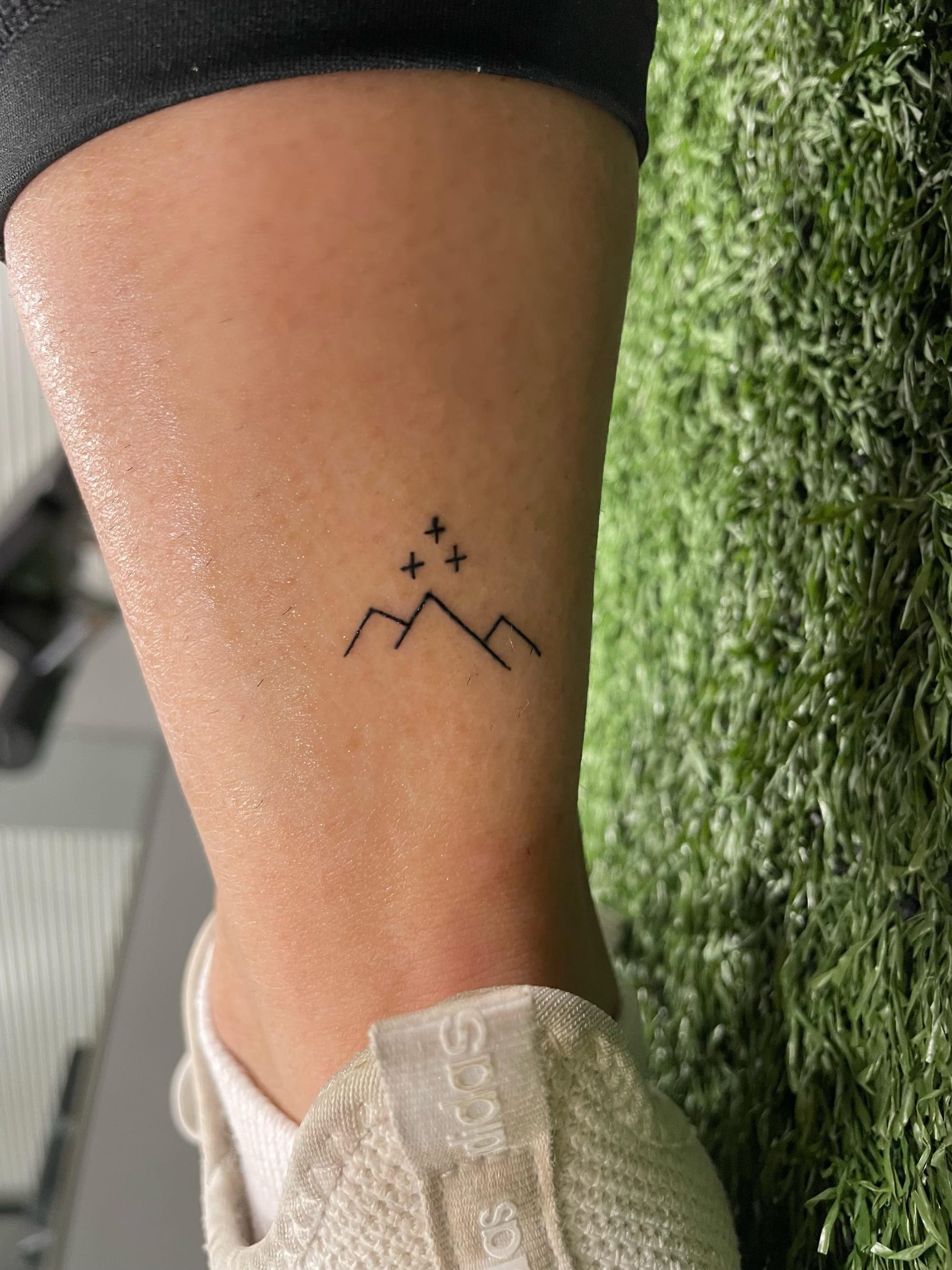 Delicate line work captures the essence of wanderlust in this minimalist mountain range tattoo, positioned perfectly on the ankle where it peeks out like a secret adventure story. Three tiny stars dance above the peaks, creating a whimsical constellation that speaks to those who find magic in both earthbound journeys and celestial dreams ⭐ The simple black ink design proves that sometimes the most meaningful tattoos whisper rather than shout.