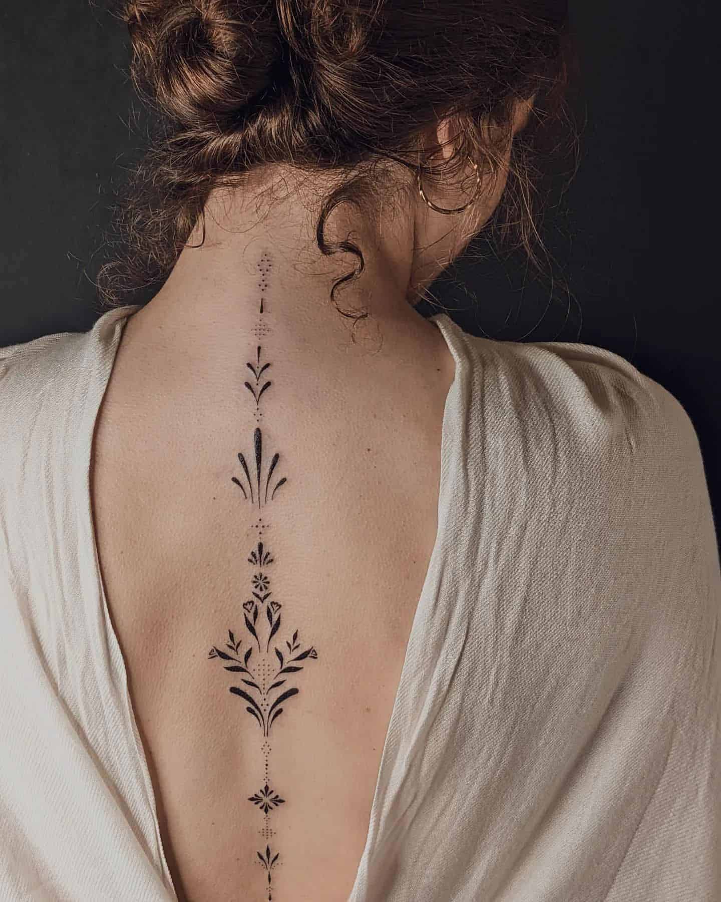 Delicate botanical elements cascade down the spine in perfect symmetry, creating a living garden that follows the natural curve of the back. The intricate linework features flowing leaves, geometric accents, and dotwork details that transform the spine into an elegant vertical canvas 🌿 This sophisticated piece proves that minimalist tattoos can make the most powerful statement.