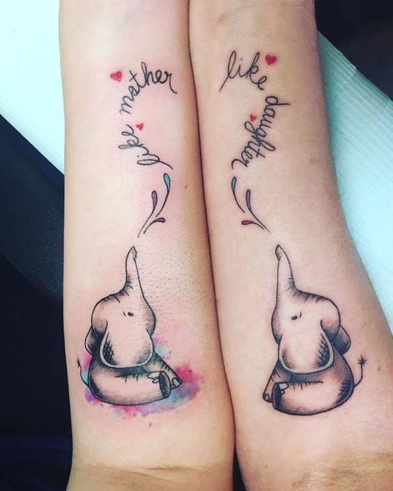Matching elephant tattoos that celebrate the unbreakable bond between mother and daughter! 🐘 The adorable baby elephants reach toward each other with their trunks while delicate script and tiny hearts float above, creating a heartwarming tribute to family love. The soft watercolor splashes beneath each elephant add a dreamy artistic touch to this meaningful forearm piece.