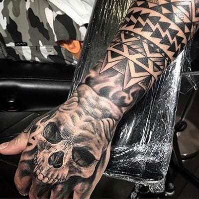 Dark artistry meets ancient symbolism in this powerful forearm piece, where a haunting skull emerges from swirling smoke beneath bold geometric patterns. The contrast between the delicate realism of the mortality motif and the sharp, tribal-inspired linework above creates a captivating visual journey from life to death. This black and gray masterpiece showcases how traditional Polynesian elements can seamlessly blend with contemporary tattoo techniques ⚫