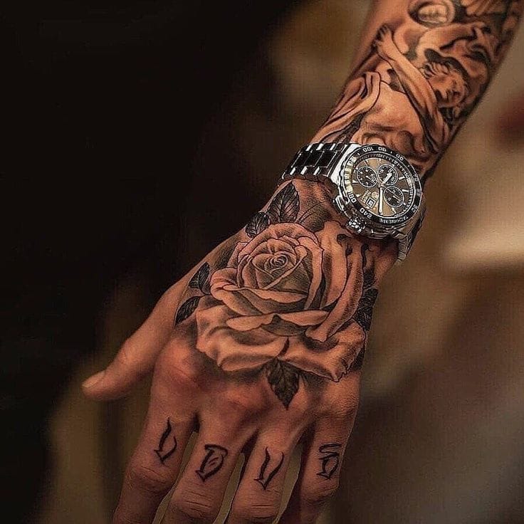 Masterful black and gray shading transforms this hand and forearm into a canvas of romantic symbolism, where detailed roses bloom alongside intricate script work. The strategic placement around a luxury timepiece creates a sophisticated contrast between organic artistry and mechanical precision, while 