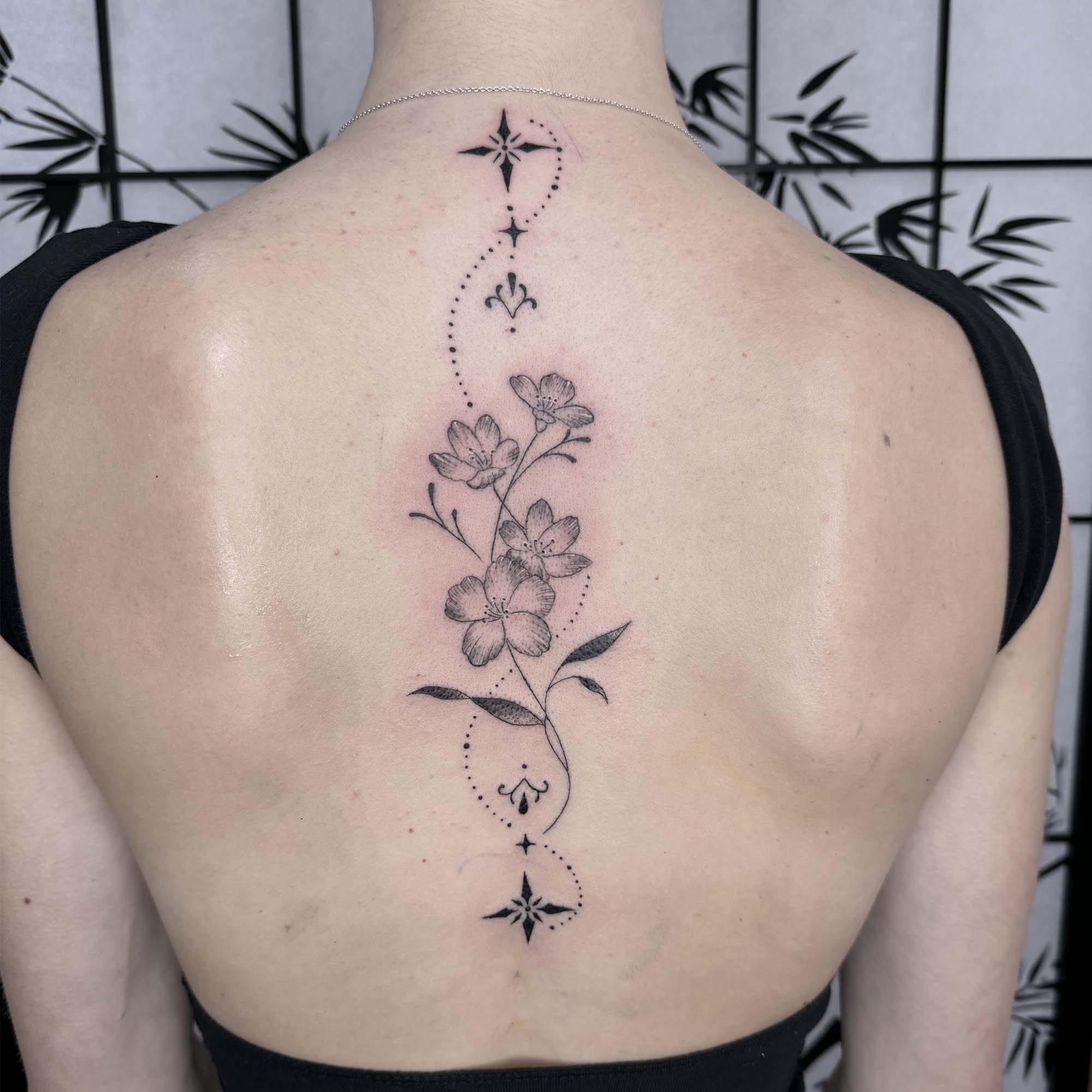 Delicate florals dance along the spine in this ethereal black and grey masterpiece, where dotted celestial elements create a mystical pathway from neck to lower back. The soft shading technique brings these blooms to life while ornate compass stars add a touch of wanderlust magic ✨