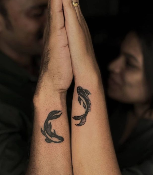 Two graceful koi fish dance across paired forearms, creating a mesmerizing visual story when hands come together in prayer position. The minimalist black ink design captures the eternal dance of yin and yang, with each fish perfectly mirroring its partner to symbolize balance and harmony. 🐟 This intimate couple's tattoo transforms a simple gesture into a meaningful display of their interconnected souls.