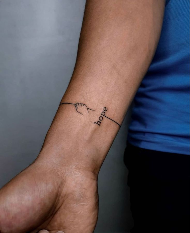 Delicate linework transforms a simple concept into profound body art with this minimalist wrist design featuring praying hands and the word