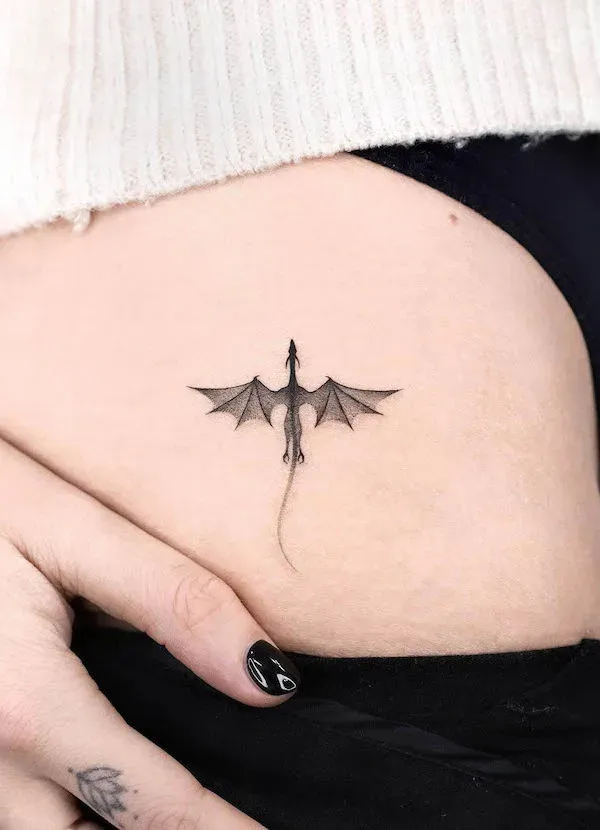 Delicate linework transforms a mythical dragon into an elegant minimalist masterpiece, perfectly positioned on the ribcage where it seems to soar across the skin. The fine black ink captures every detail of the outstretched wings and serpentine tail, creating a tattoo that's both mystical and refined ✨ A stunning example of how fantasy creatures can be reimagined through subtle, sophisticated artistry.