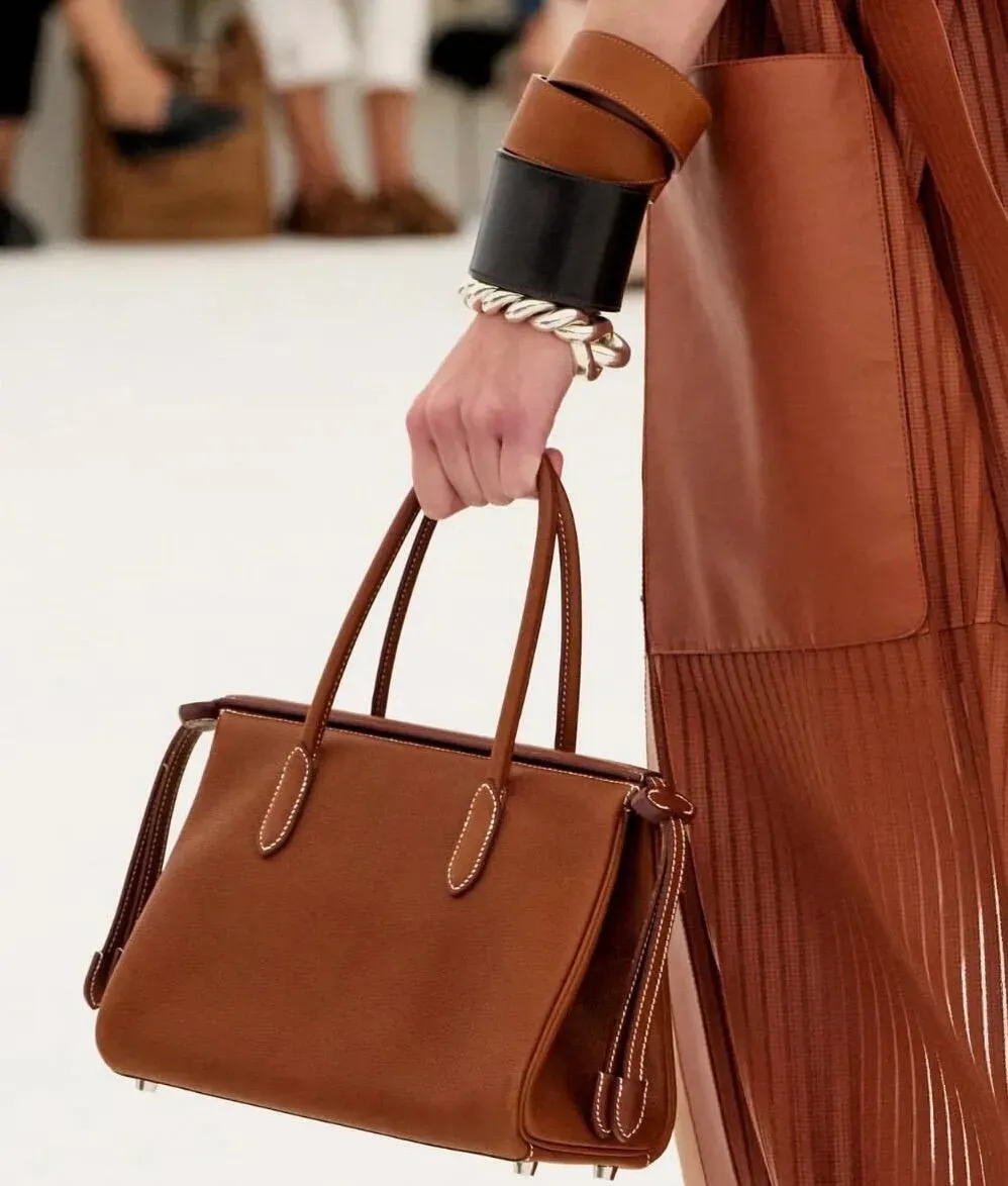 Rênes Bag（Birkin-Inspired Tote）以Birkin輪廓為靈感，採用柔軟小牛皮打造梯形袋身，前後加入加長皮革帶。