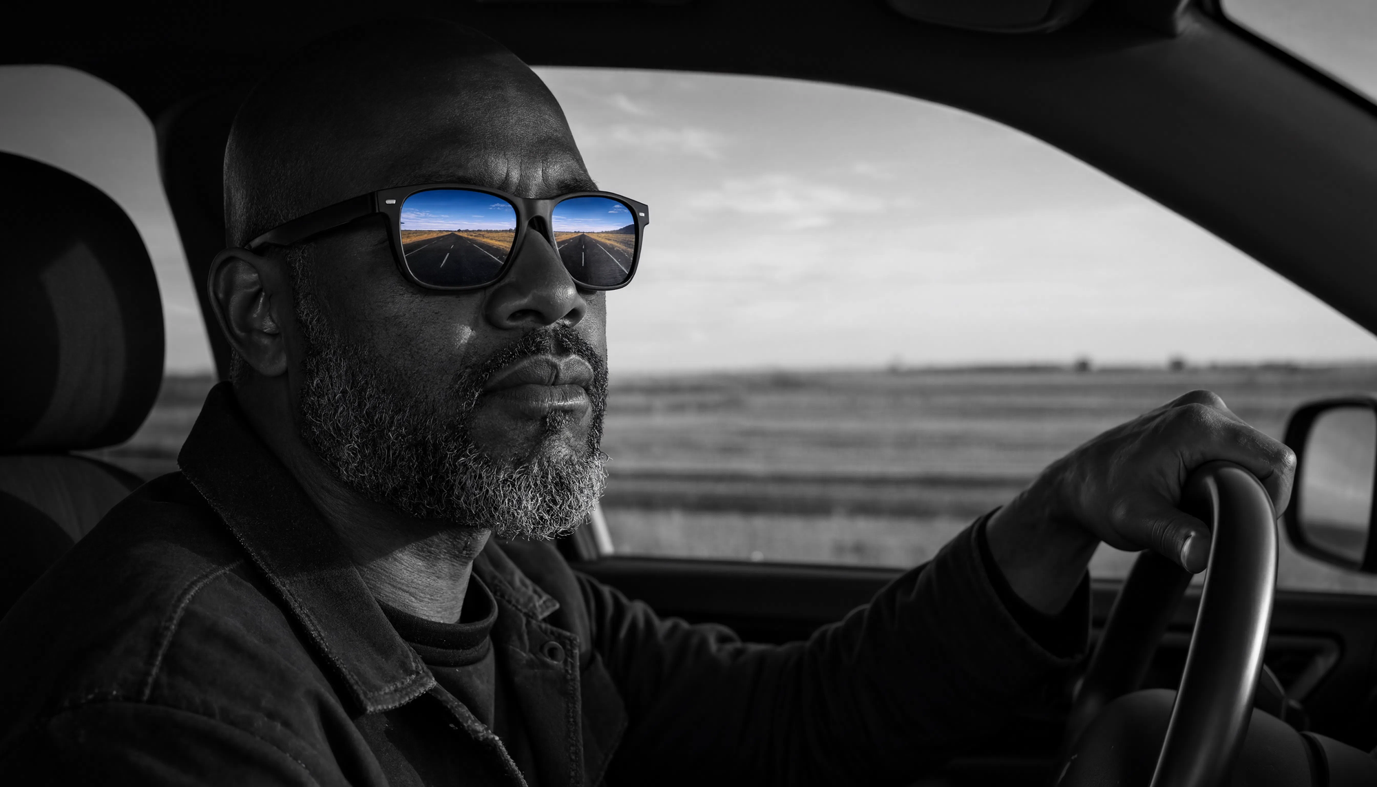 A bald Foundational Black American man with a salt-and-pepper beard drives, the car and landscape around him in black-and-white, an open empty road reflected in color in his sunglasses.