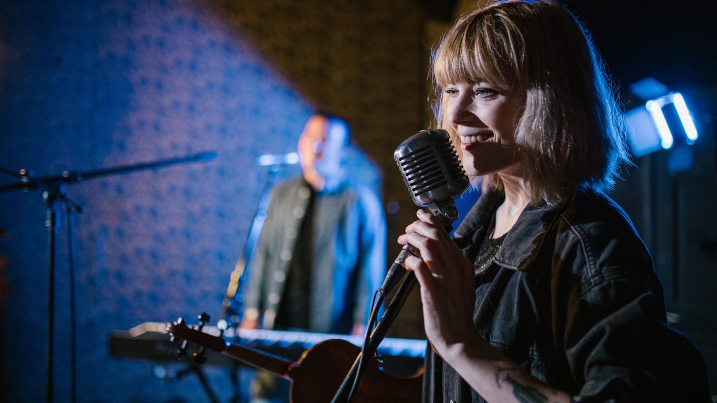 Two people on a blue-lit stage: one in close focus, smiling into a ribbon mic and holding a violin, the other blurry in the background, standing at a key board.