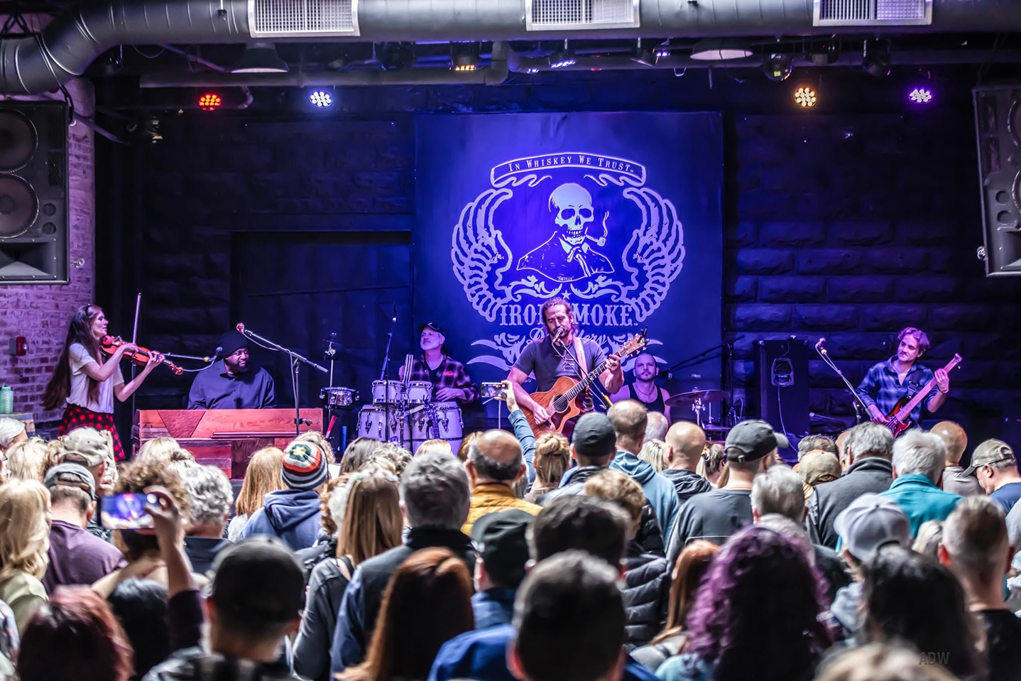 The Adam Ezra band live on stage, playing: violin, keyboard, percussion set, drum kit, bass, and guitar. 